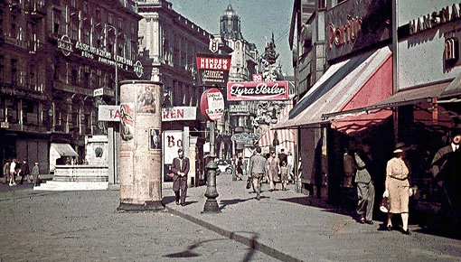 Auf der nachkolorierten Fotografie sieht man Menschen vor Schaufenstern und beim Bummeln.