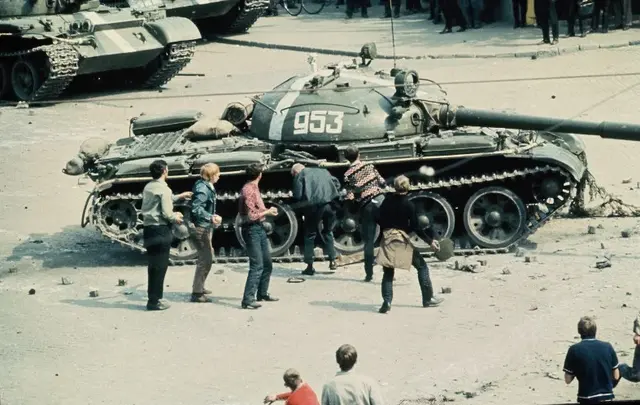 Einige junge Männer, teilweise Steine werfend, neben einem sowjetischen Panzer auf der Straße. Im Hintergrund sind die Ketten weiterer Panzer sowie Füße von Schaulustigen zu sehen.