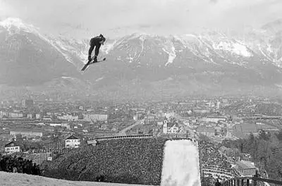 Blick vom Berg Isel auf die Stadt während des Skisprungbewerbs. Ein kleiner Skispringer ist vor dem Bergpanorama und dem Auslauf zu sehen.