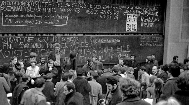 AudiMax der Wiener Universität. Eine Menge Studenten drängen sich vor der Tafel des Hörsaales zusammen.