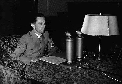 Goebbels bei einer Rundfunkansprache zum bevorstehenden 50. Geburtstag von Adolf Hitler am 19. April 1939. Goebbels in Uniform vor einem Manuskript, zwei Mikrofonen und einer Lampe sitzend.