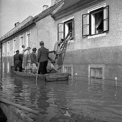 Feuerwehrleute sind mit Zillen unterwegs und betreuen die Flutopfer, an die sie amerikanische CARE-Pakete verteilen. Linz, 14. Juli 1954. Eine Frau übernimmt ein Paket und klettert auf einer Leiter durchs Fenster der Zille entgegen.