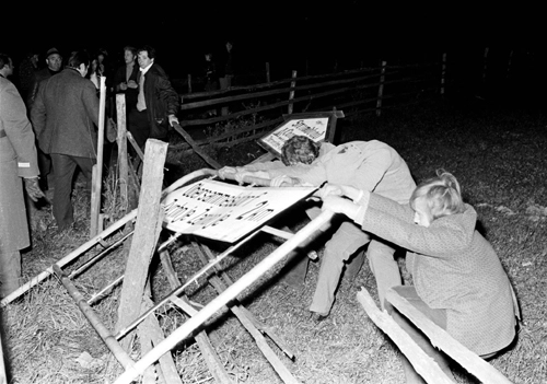 Zwei Männer zerren an dem schon schräg liegenden zweisprachigen Hinweisschild.