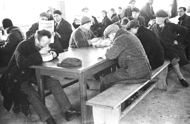 Arbeitslose bekommen eine warme Mahlzeit. Steyr, 1932. Männer in dicken Mänteln sitzen an Tischen, essen, lesen Zeitung und schauen teilweise in die Kamera.