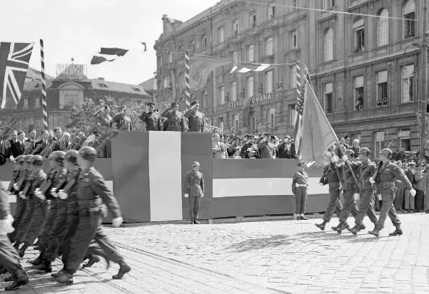 Salutierende Kommandeure auf rot-weiss-rot verkleideten Tribünen nehmen ein Militärparade ab. Hier sind vier US-amerikanische Soldaten mit Stahlhelmen - zwei mit geschulterten Gewehren und zwei mit Flaggen zu erkennen. Ganz links marschieren Soldaten aus dem Bild.