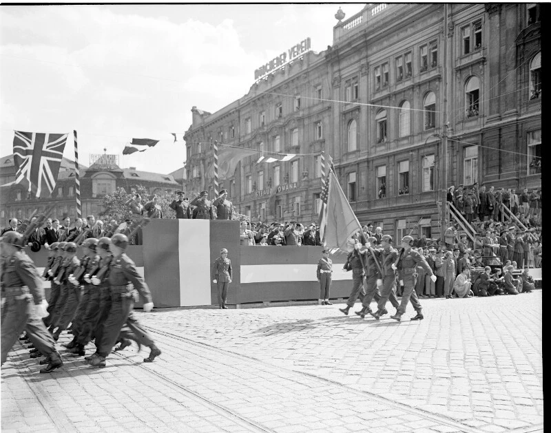 Salutierende Kommandeure auf rot-weiss-rot verkleideten Tribünen nehmen ein Militärparade ab. Hier sind vier US-amerikanische Soldaten mit Stahlhelmen - zwei mit geschulterten Gewehren und zwei mit Flaggen zu erkennen. Ganz links marschieren Soldaten aus dem Bild.