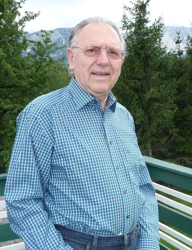 Gerhard Jagschitz als älterer Herr im blauen Hemd blickt in die Kamera. Im Hinterbrund sind ein Geländer, Wald und Berge sichtbar.