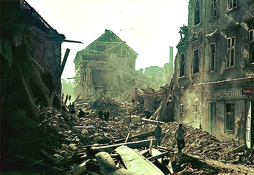 Blick in eine zerstörte Straße in Wien nach einem Bombenangriff mit Schutt auf den Straßen, wo schon Leute schauen, was passiert ist und einige Häuser halb zerstört sind. (Ein seltenes Farbfoto).