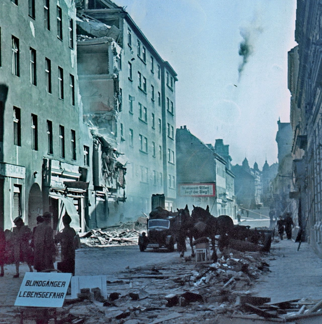 Straße in Wien nach einem Bombenangriff 1945. Im Vordergrund sieht man einen Warnhinweis auf eine nicht detonierte Bombe: "Blindgänger Lebensgefahr". Im Hintergrund ist ein rot eingerahmtes Propagandaplakat mit der Aufschrift: "In unserem Willen liegt der Sieg!" zu erkennen.