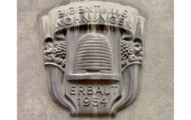 Stuck-Logo an einer Hauswand. Ein Bienenkorb. Darüber: Eigentumswohnungen. Erbaut 1954".