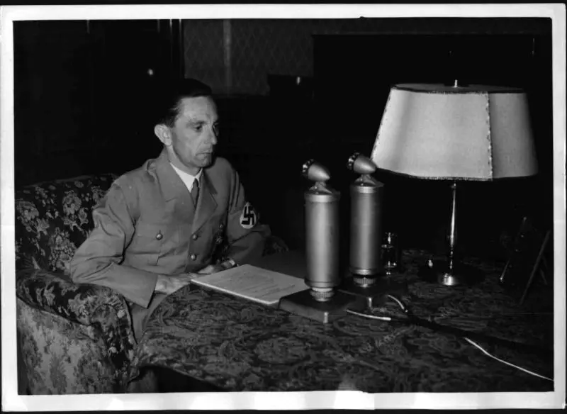 Goebbels bei einer Rundfunkansprache zum bevorstehenden 50. Geburtstag von Adolf Hitler am 19. April 1939. Goebbels in Uniform vor einem Manuskript, zwei Mikrofonen und einer Lampe sitzend.