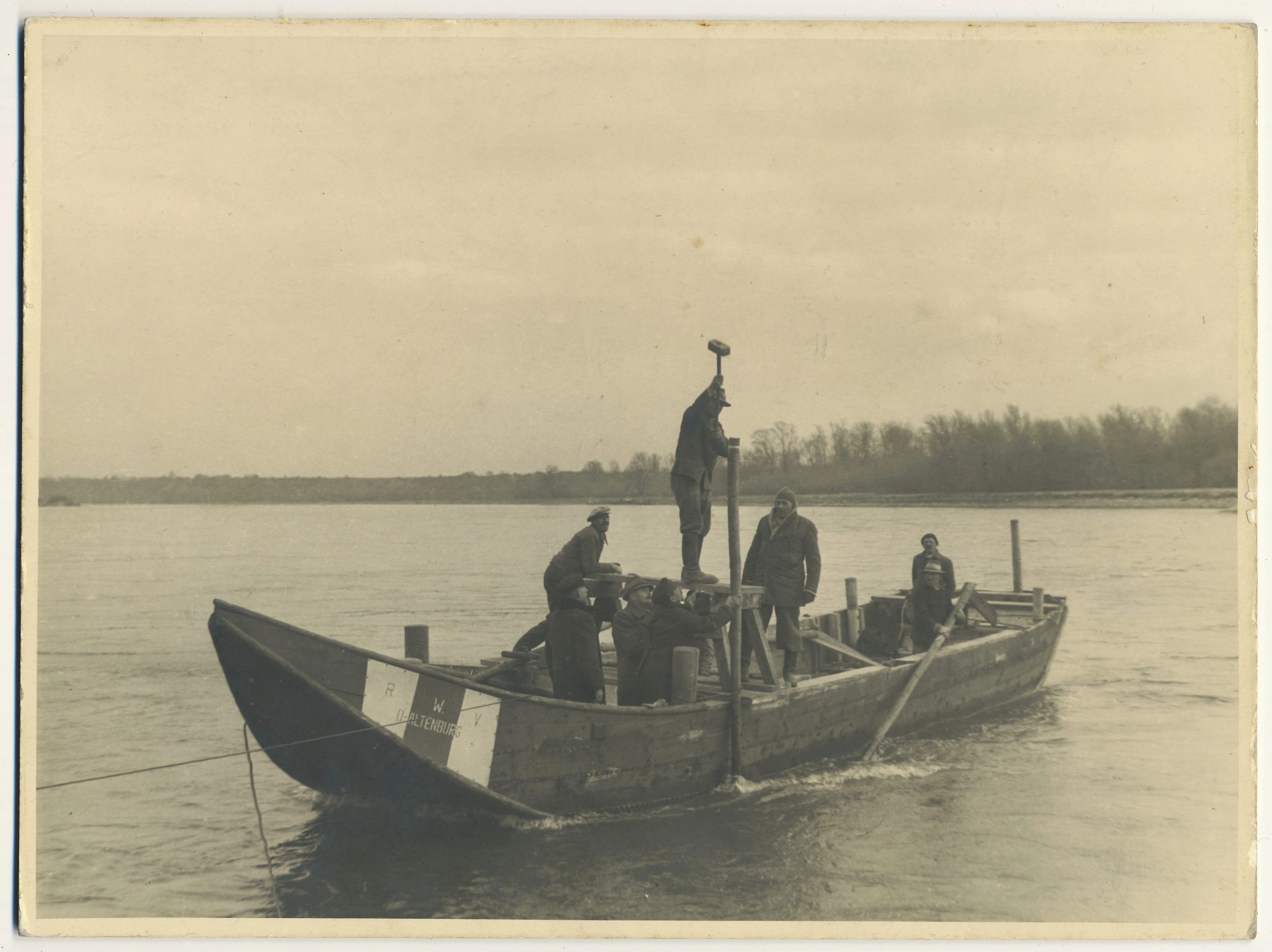 Acht Arbeiter stehen auf einem Boot in der zufrierenden Donau. Ein Arbeiter schlägt mit einem schweren Hammer einen dicken Pfosten ein. Ein zweiter Arbeiter hält den Pfosten gerade.