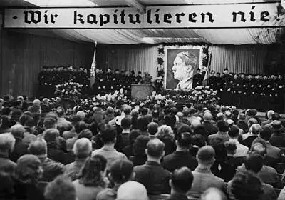 Bei einem Betriebsappell spricht Reichsorganisationsleiter Robert Ley in einem großen Berliner Rüstungsbetrieb, am 5. Februar 1941. Gefüllter Saal und volle Tribüne. Auf dieser ein Hitlerportrait und darüber ein Transparent: "Wir kapitulieren nie!"