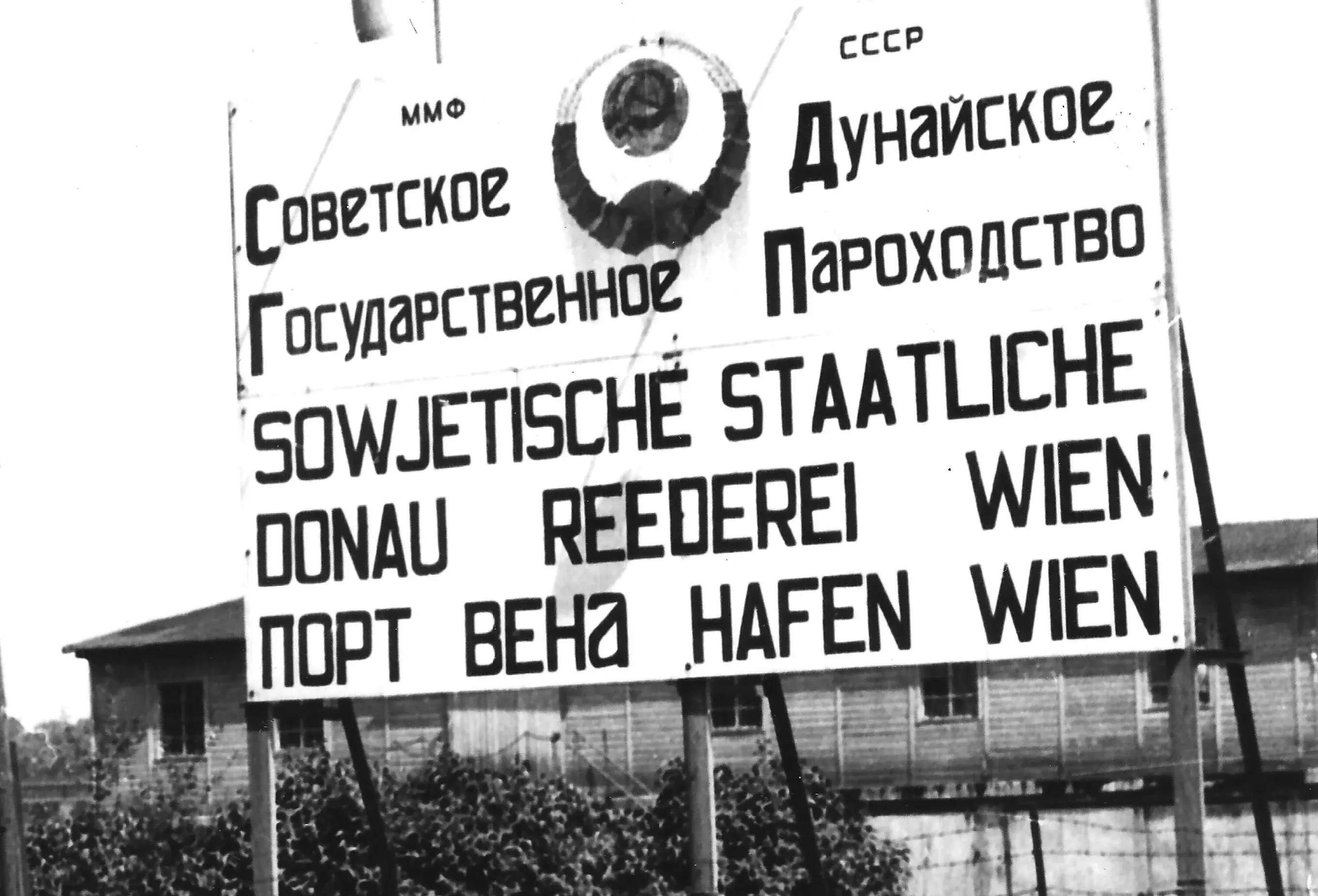 Schild mit Aufschrift Sowjetische staatliche Donau Reederei Wien - Hafen Wien und russische Übersetzung in cyrilischen Buchstaben. Im Hintergrund Zäune und Schinen im Vordergrund. Das Schild daneben ist schlecht erkennbar.