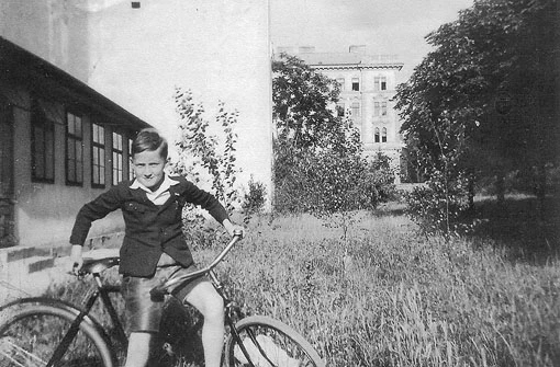 Günther Schifter als Kind steht mit einem Herrenfahrrad auf einer hohen Wiese vor einem Haus.